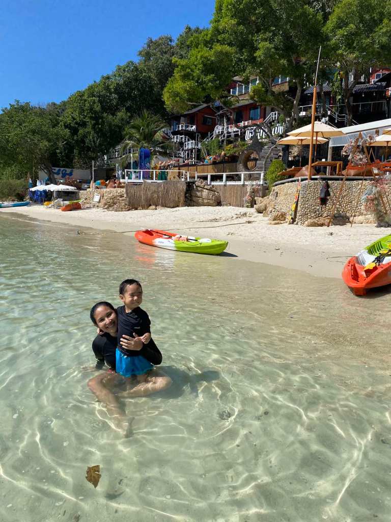 Mom and son smiling at the beach with clear waters and kayaks at the back.