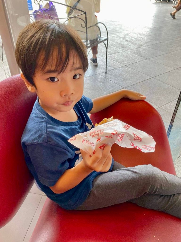 A boy sitting cross legged on a red chair, eating a chicken burger wrapped with a sandwich wrapper