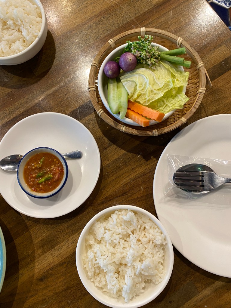 Wooden table laden with plates and utensils, a bowl of shrimp paste, another with slices of fresh cabbages, carrots, cucumbers, beans, and eggplants, and a small cup of white rice.