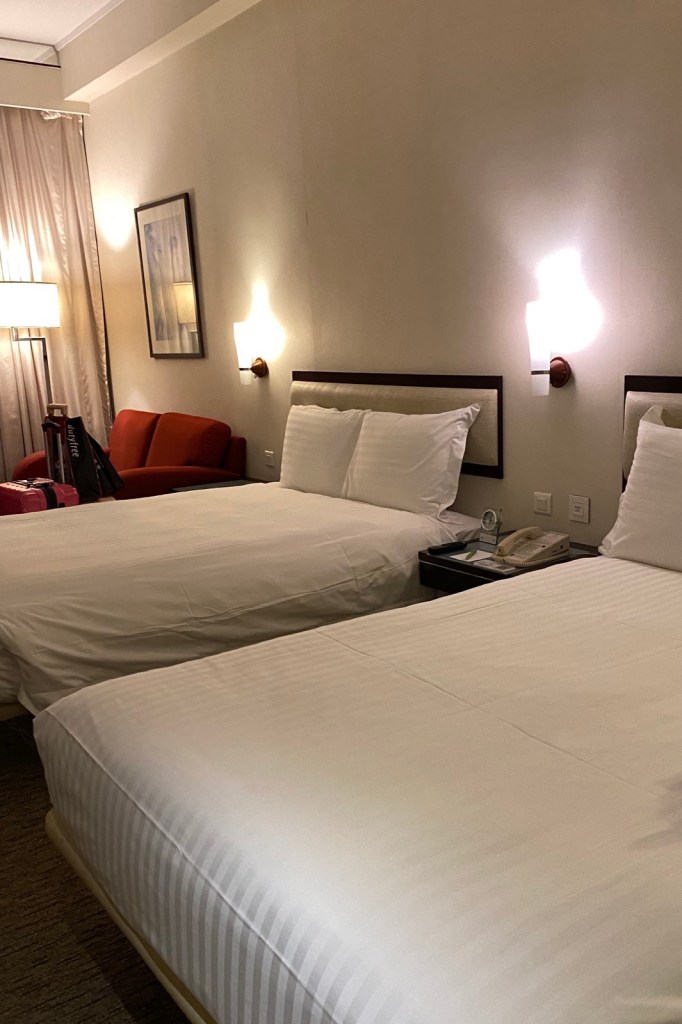 A hotel room with two beds made with white, striped linens. Between the beds is a nightstand, and in the background, there is a red sofa and a floor lamp.