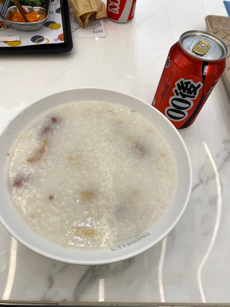 A top-down view of a white bowl filled with congee (rice porridge) next to a red can of Coca-Cola, which has Asian characters, on a white marble table.