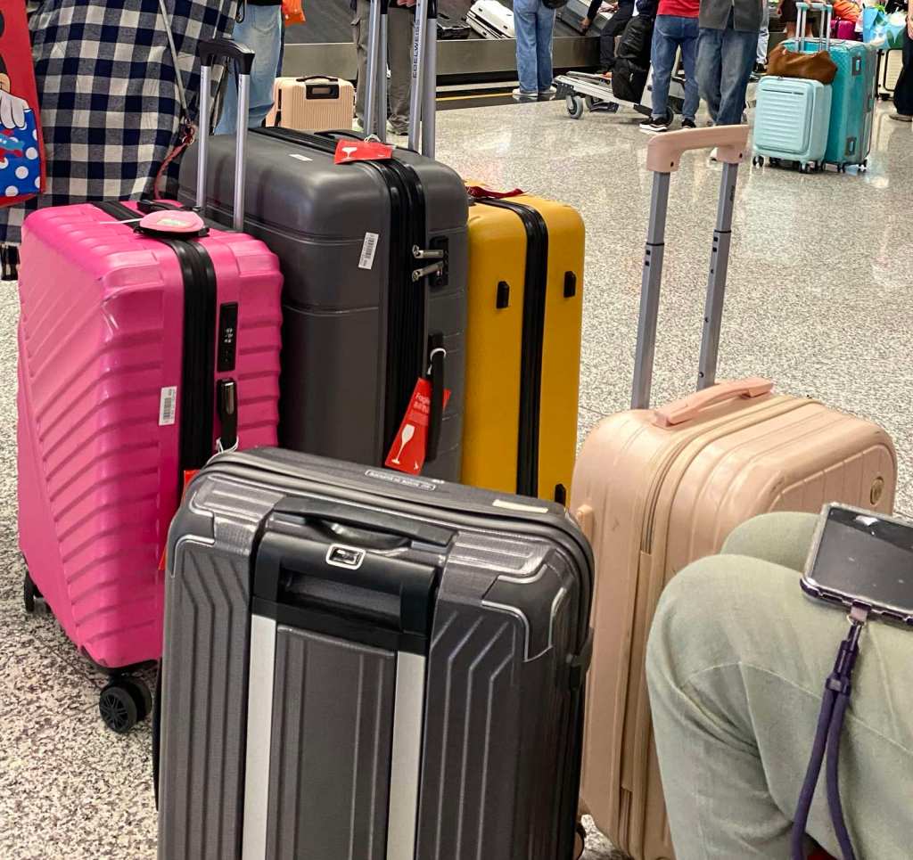 A cluster of five roller suitcases (pink, grey, yellow, and beige) on the floor of an airport baggage claim, with red "fragile" tags on two of the bags.