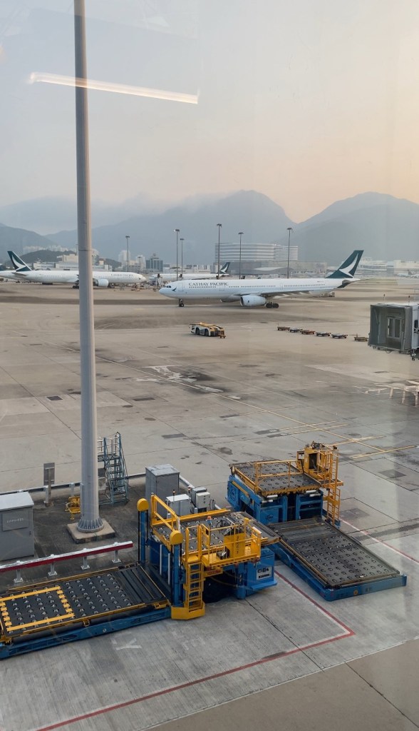 A view from an airport terminal window overlooking the tarmac, showing several Cathay Pacific airplanes parked, with yellow and blue baggage handling equipment in the foreground and mountains visible in the hazy background.