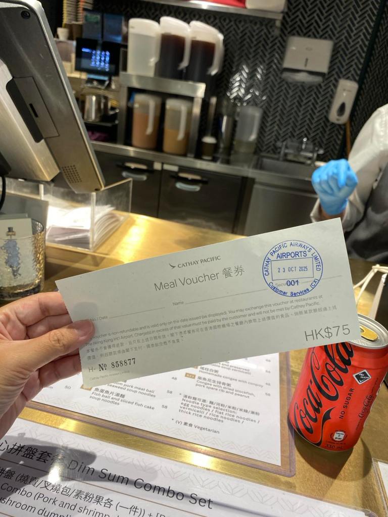 A hand holds up a Cathay Pacific meal voucher valued at "HK$75" and stamped "23 OCT 2025" in front of an airport restaurant counter.